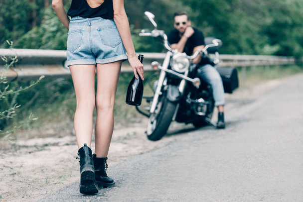 обрізаний вид молодої жінки, що йде по дорозі з пляшкою алкоголю біля хлопця на чорному мотоциклі
 - Фото, зображення