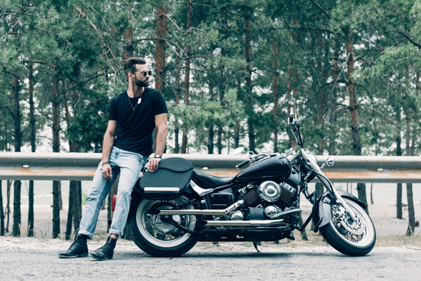 guapo motociclista en gafas de sol de pie cerca de motocicleta negro en la carretera cerca de bosque verde
 - Foto, Imagen
