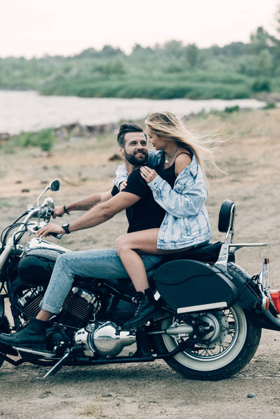 smiling young couple of bikers hugging on black motorcycle at sandy beach - Zdjęcie, obraz