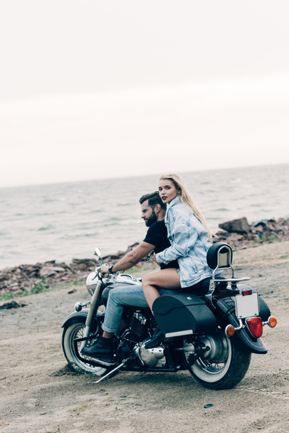 νεαρό ζευγάρι μοτοσικλετιστών που κάθονται σε μαύρη μοτοσικλέτα στην παραλία κοντά στο ποτάμι - Φωτογραφία, εικόνα