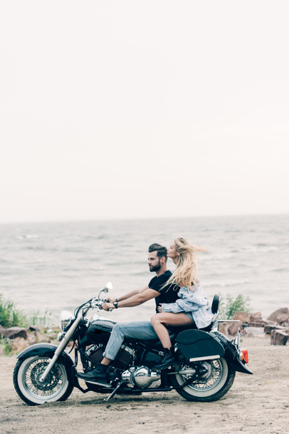 Widok z boku młodej pary motocyklistów jeżdżąc czarnym motocyklem na piaszczystej plaży w pobliżu rzeki - Zdjęcie, obraz