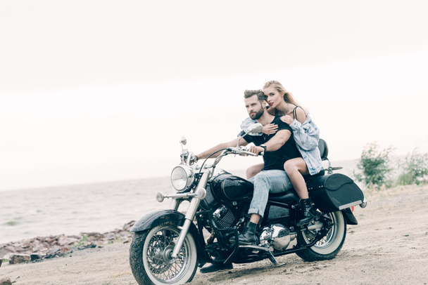 Młoda para rowerzystów jazda na czarnym motocyklu i obejmując na piaszczystej plaży - Zdjęcie, obraz