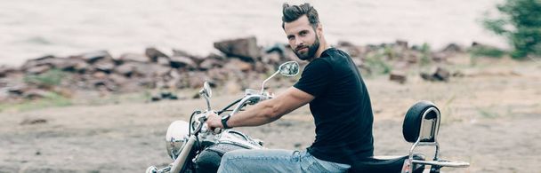 handsome bearded motorcyclist on black motorcycle at sandy beach near sea, panoramic shot - Photo, Image