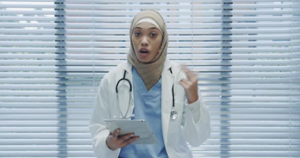 Waist up lockdown shot of young mixed race female doctor wearing hajib using tablet, gesturing and talking to camera in hospital  - Materiał filmowy, wideo