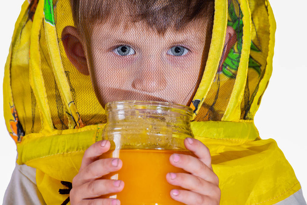 Un niño con un traje amarillo protector de abejas sosteniendo un tarro de miel en sus manos. Foto aislada
. - Foto, Imagen