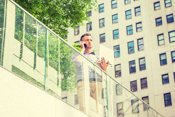 Camino al éxito. Joven estudiante americano guapo estudiando en Nueva York, de pie junto a la barandilla de vidrio en el distrito de negocios con edificios altos, árboles verdes, trabajando en el ordenador portátil, hablando por teléfono celular
 - Foto, Imagen