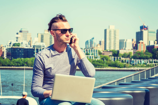 Young American man traveling, working in New York, wearing gray, long sleeve T shirt, sunglasses, sitting by river, working on laptop computer, talking on cell phone. Brooklyn buildings on background - Zdjęcie, obraz