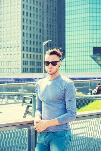 Jonge knappe man reist in New York City, het dragen van Gray, lange mouw T shirt, zonnebril, staande op Park in zakenwijk met hoge gebouw, onder de zon, rondkijken, ontspannen, denken - Foto, afbeelding