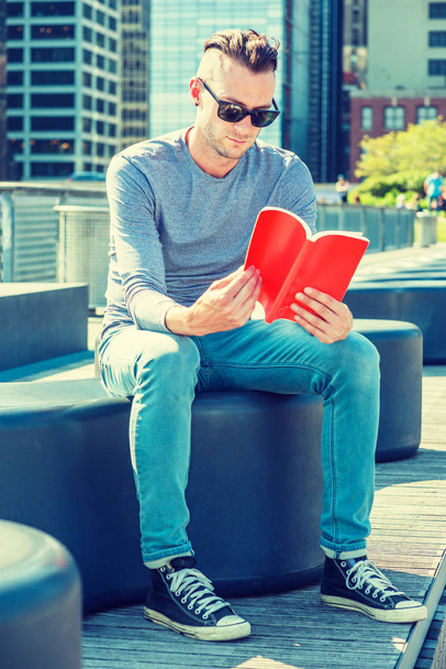 私は本を読むのが大好きです。灰色の長袖Tシャツ、青いジーンズ、黒いスニーカー、サングラスを着た若いアメリカ人男性、ニューヨーク市のビジネス地区の公園に座って、太陽の下で、赤い本を読んで - 写真・画像