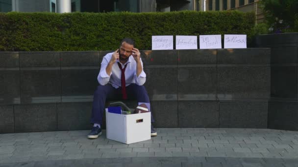 Hombre de negocios despedido sentado frustrado y molesto en la calle cerca del edificio de oficinas con caja de sus pertenencias. Perdió el trabajo. Alrededor de los envases de cartón descompuestos etiquetados, despedidos, desempleados, necesitan trabajo
 - Imágenes, Vídeo