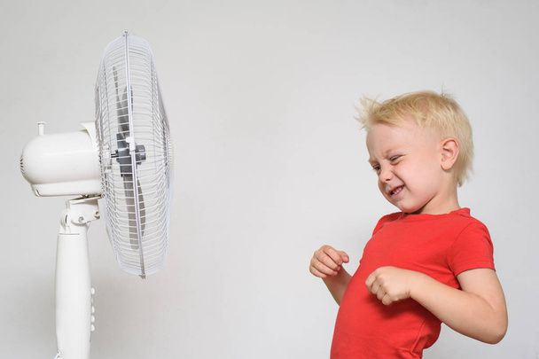 Kleine blonde jongen in een rood T-shirt maakt een gezicht in de buurt van de ventilator. Zomer concept - Foto, afbeelding