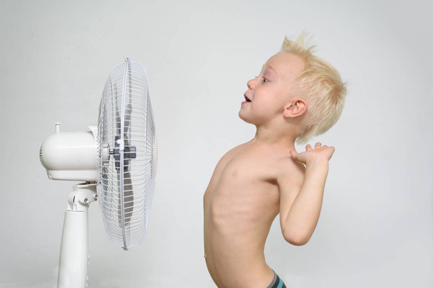 Удивленный маленький блондин с обнаженным туловищем стоит рядом с вентилятором. Летняя концепция
 - Фото, изображение