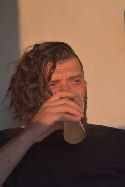 Όμορφος νεαρός άντρας με μακριά σγουρά μαλλιά και μούσι. Άνθρωπος βάναυση μελαχρινή σε ένα μαύρο T-shirt και με ένα μοδάτο κούρεμα! Ο τύπος πίνει μπύρα.. - Φωτογραφία, εικόνα