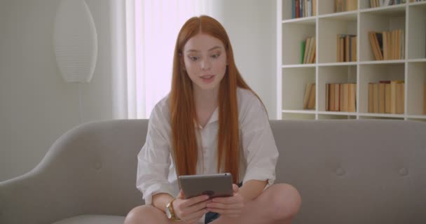 Closeup portrait of young pretty redhead female having a video call on the tablet talking with excitement sitting on the sofa in a cozy apartment - Materiał filmowy, wideo