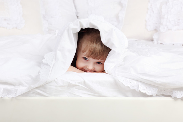 Little beautiful girl looks out from under warm blanket on bed. - Foto, afbeelding