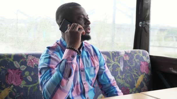 Afro americano uomo chiama e parla smartphone seduto in caffè in attesa del suo ordine
. - Filmati, video
