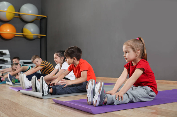 Lindos niños pequeños sentados en el suelo y haciendo ejercicio físico en el gimnasio de la escuela. Estilo de vida saludable
 - Foto, imagen