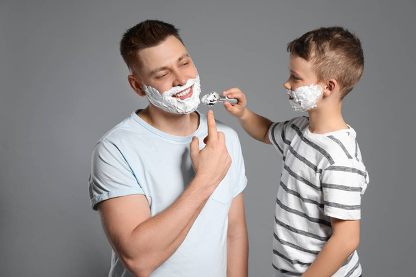Son applying shaving foam on dad's face, grey background - Фото, изображение