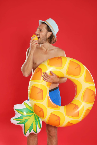 Attractive young man in swimwear with pineapple inflatable ring eating lemon on red background - Фото, изображение