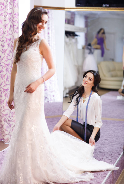 Frau probiert Hochzeitskleid im Geschäft mit weiblicher Assistentin an. - Foto, Bild