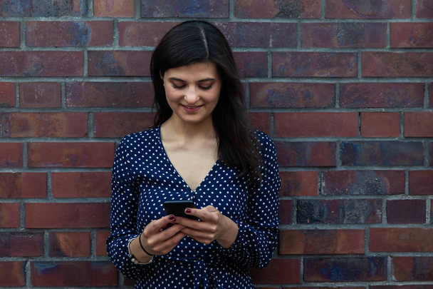 Smiling young woman using phone while leaning against brick wall - Photo, Image