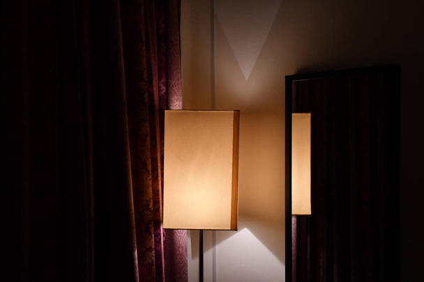 Geometrie van eenvoudige lampenkap weerspiegeld in spiegel op de muur van de kamer met donker rode sierlijke gordijnen. Gouden licht en driehoek schaduwen in de hoek van de kamer in de duisternis. Minimale interieur decoratie. - Foto, afbeelding