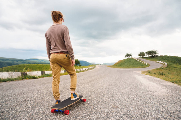 Young stylish man with long hair gathered in a ponytail and in sunglasses stands with a longboard on a country asphalt road in the mountains on the background of epic rocks - Foto, Imagen
