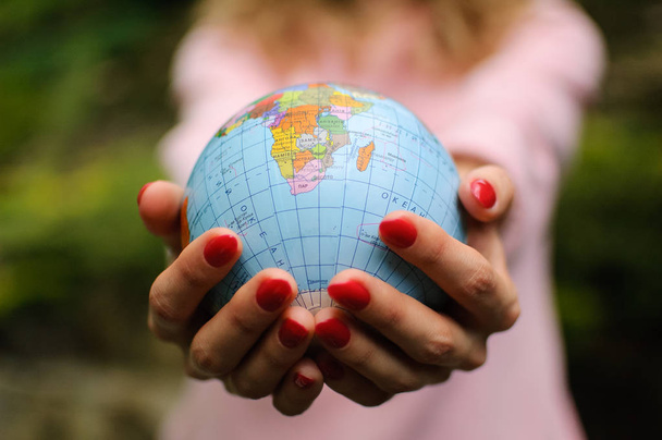 The girl in a pink sweater with red manicure holds a small globe with geografical names in Ukrainian cyrillic letters on it. African continent and Ocean are visible. - Foto, imagen