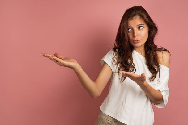 Young brunette points at the left space at the picture with a question in her eyes, pink background - Photo, image