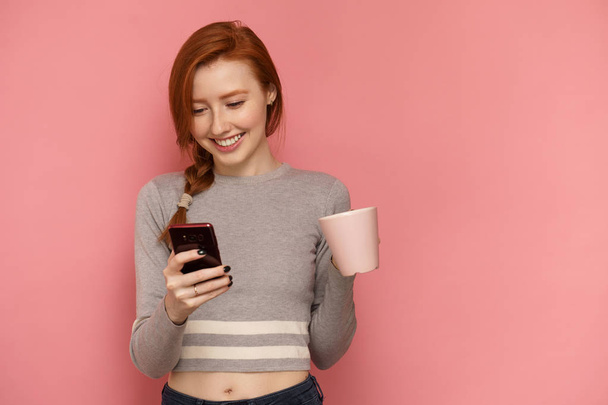 Red-haired girl holds a cup in one hand looking at her smartphone in other hand and smiling - Fotoğraf, Görsel