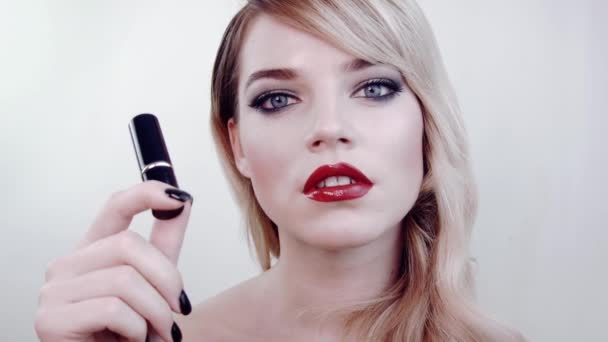 Beautiful girl holding closed black lipstick tube from side of face.Choose lipstick.She thinks about buying.Look at camera.In studio on white background.Beauty salon. Hair in retro style,bright makeup - Кадри, відео
