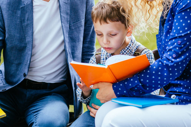 kihloissa oleva lapsi poika istuu äidin ja isän välillä ja lukee mielenkiintoista kirjaa ulkona
. - Valokuva, kuva