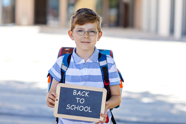 Onnellinen pikkupoika, jolla on reppu, olkalaukku ja lasit. Koulupoika on matkalla kouluun. Terve suloinen lapsi ulkona liitupöydällä Hei hei koulu. Takaisin kouluun käsite
 - Valokuva, kuva