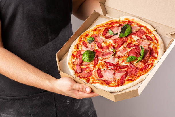Bir site veya menü için reklam fotoğrafı. Koyu renk önlüklü genç bir adamın elinde bir kutuda jambon ve salamla pizza. parlak arka plan. - Fotoğraf, Görsel