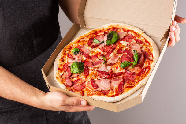 Advertising photo for a site or menu. Pizza with ham and salami in a box in the hands of a young man in dark apron. bright background. - Zdjęcie, obraz
