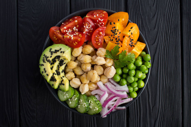 Vegane Poke-Schüssel mit Kichererbsen und Gemüse in der dunklen Schüssel in der Mitte des schwarzen Holzhintergrundes.. - Foto, Bild