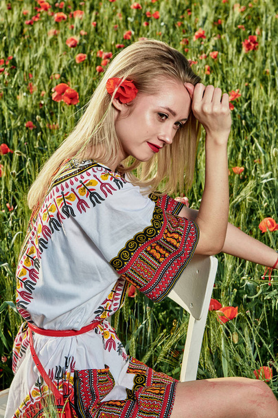 Long-haired blonde young woman in a white short dress on a field of green wheat and wild poppies. - Φωτογραφία, εικόνα
