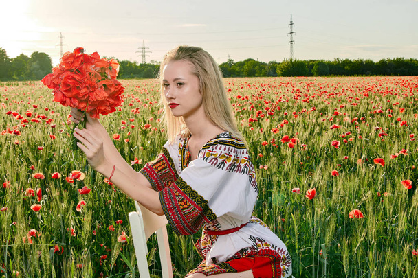 Long-haired blonde young woman in a white short dress on a field of green wheat and wild poppies. - Photo, Image