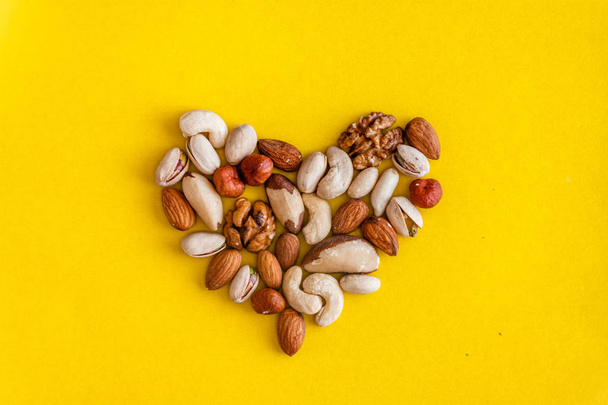 A variety of nuts are laid out in the shape of a heart on a yellow background. Flat lay. View from above. Copy space. The concept of health care, cardio, proper nutrition, healthy fats - Zdjęcie, obraz