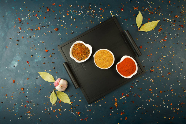 SPICES IN BOX ON A DARK BACKGROUND WITH THE GARLIC AND BAY LEAF. BRIGHT SPICES DISHES STANDING ON A WOODEN TRAY WITH HANDLES ON A DARK TEXTURED BACKGROUND WITH THE GARLIC AND BAY LEAF. - Fotoğraf, Görsel