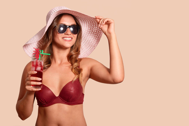 Obraz veselého, dobře vypadající turistiky s slaměným kloboukem, červeným plaveckým oblečením a slunečními brýlemi, který si hledí stranou, vychutnával si letní prázdniny a sklenici studeného koktejlu. Kopírovat prostor pro reklamu. - Fotografie, Obrázek