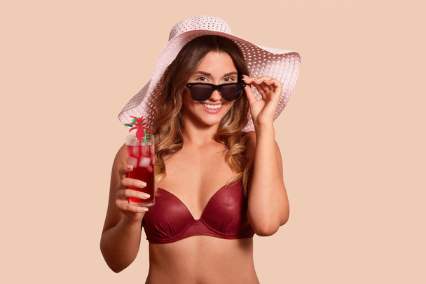 Indoor studio shot of beautiful magnetic model posing isolated over beige background, holding cold summer drink in one hand, touching her eyewear with fingers, wearing accessories. Vacation concept. - Valokuva, kuva