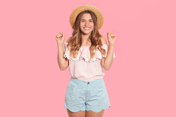 Studio shot of cheerful charming female wearing straw hat, light blouse and shorts, raising her hands, being in high spirits, looking directly at camera, having wavy hair. People and emotions concept. - Zdjęcie, obraz