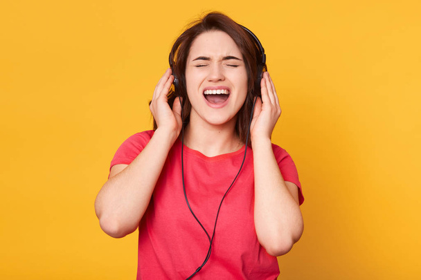 Image of attractive energetic brunette wearing casual red t shirt, having headphones, listening to music, singing, enjoying spending her spare time alone, closing eyes, opening mouth widely. - Foto, afbeelding