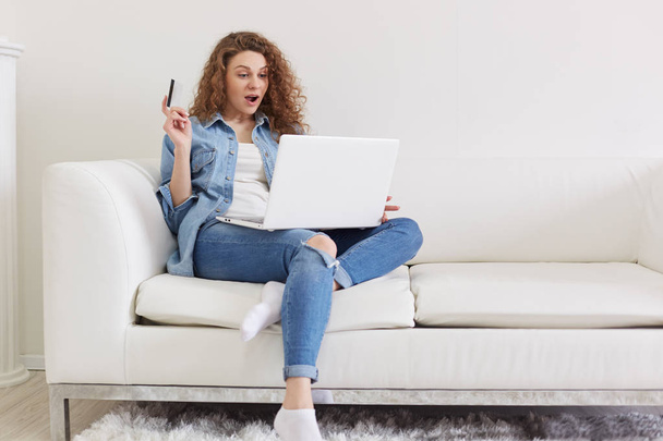 Indoor shot of shocked emotional lady with curly hair sitting in light living room, raising one hand, holding credit card, using her laptop, doing purchases on net, being fond of shopping online. - Foto, Imagen