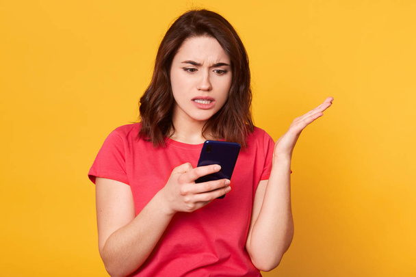 Impressed emotional young lady raising her arm with indignation, having unpleasant facial expression, holding smartphone, standing isolated over yellow background in studio. Technology concept. - Fotografie, Obrázek
