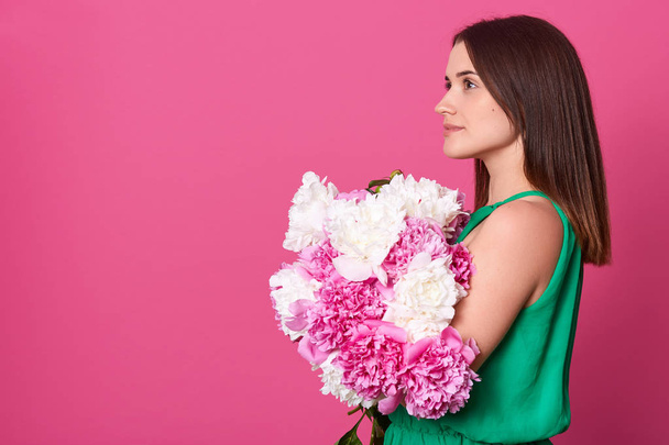 Profile of thoughtful attractive European woman looking straight forward, holding flowers, standing isolated over pink background in studio, wearing green dress. Copyspace for advertisement. - Photo, Image