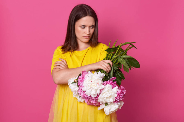 Upset disappointed young woman standing with folded arms, holding flowers in one hand, looking aside, having unpleasant facial expression, having short dark hair, wearing bright yellow dress. - Foto, afbeelding