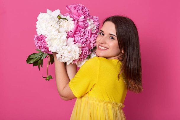 Portrait of cheerful happy cute model standing isolated over pink background in studio, smelling and putting close to face white and peonies, going to important event. People and emotions concept. - Foto, Bild