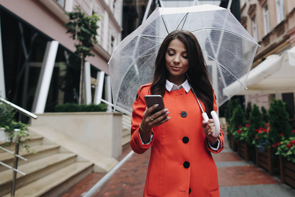 Beautiful brunette woman with umbrella in red coat dialing numbe - Фото, зображення